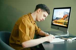 High school male student sitting in front of a computer and writing in a notebook.