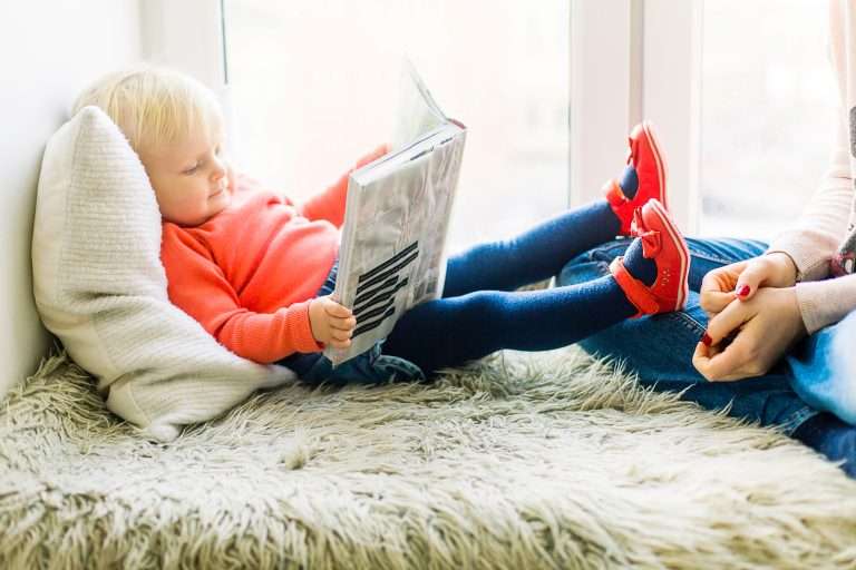 Toddler laying on a bed reading a book with mother sitting nearby.