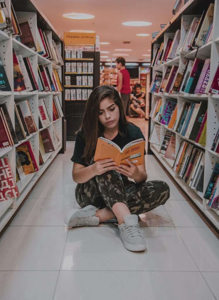 Girl sitting on the floor between two book-filled shelves reading a book.