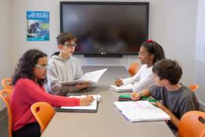 4 students sitting at a table discussing and writing in notebooks.