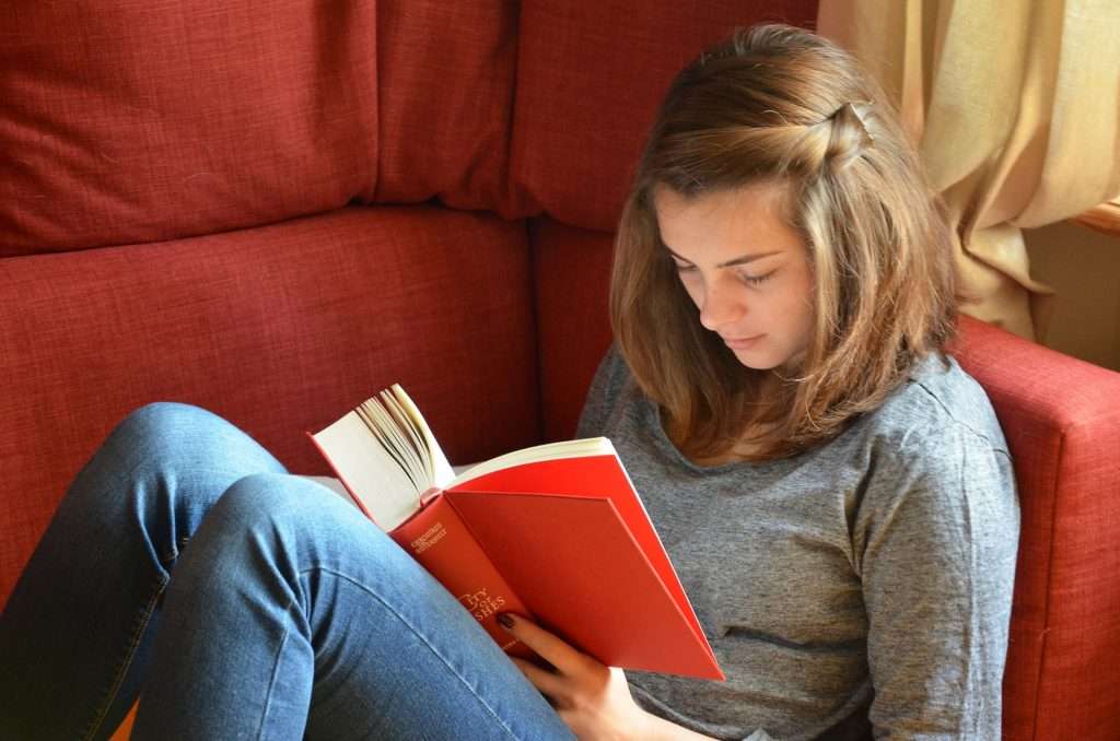 Adolescent female sitting on a couch reading a book.